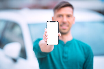 Smartphone mockup, blank screen. Young smiling man standing near white car outdoors and showing or holding his phone with blank space