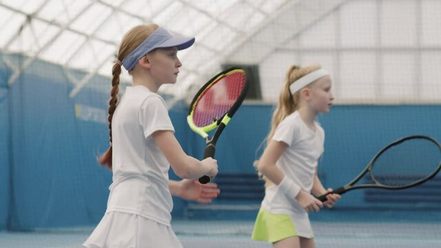 Slow-motion medium shot of two young caucasian girls in white tennis uniform hitting tennis ball during indoor training on court