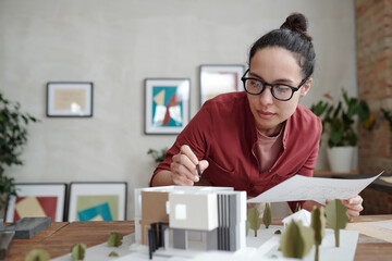 Young elegant female architect with paper and pen bending over model of new house and yard and pointing at roof while working over project
