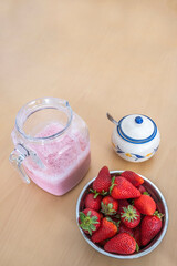 bowl full of strawberries and a jar with freshly made strawberry milkshake, a sugar bowl, ingredients for a natural strawberry smoothie, on a light wooden table, copy space