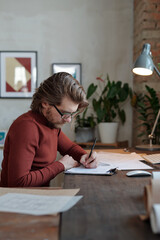 Fototapeta na wymiar Young serious male architect in eyeglasses and casualwear concentrating on making sketch of new construction while sitting by table in office
