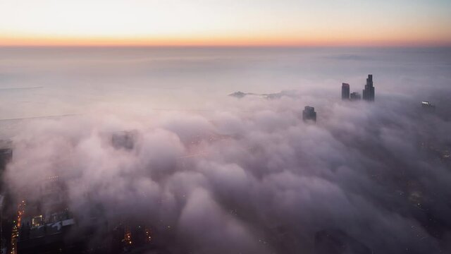 Beautiful Chicago time lapse aerial of heavy low fog rolling into the city at sunrise towards Lake Michigan as the weather begins to warm up and the sun casts pink and orange hues on the clouds below.