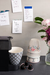 Many reminder notes posted in a gray wooden wall next to a coffee machine, decaf capsules and three cups in a pile.
