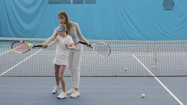 Slow-motion full shot of young female tennis coach helping little girl to hold tennis racket properly training together on court indoors