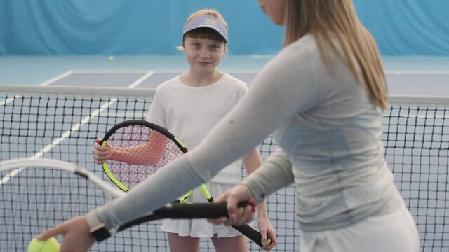 Slow-motion medium shot of cheerful little girl learning to play tennis on court indoors while female coach getting ready to throw tennis ball with racket