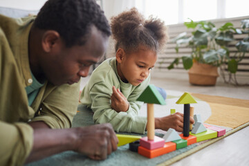 Portrait of African-American man playing with daughter while lying on floor in cozy home interior,...