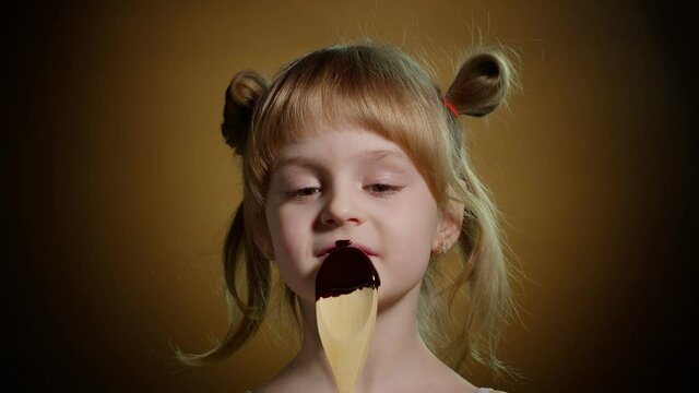Close-up portrait of teen child kid eating, licking melted chocolate sweet candy syrup from wooden spoon. Young playful blond girl with two ponytails smiling on dark brown background. Satisfied face