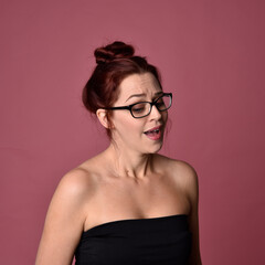 Close up portrait of a red haired woman with expressive facial features and glasses on a pink studio background.