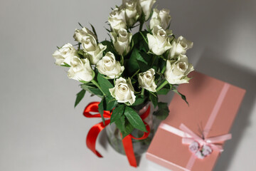 White tea roses stand in a glass vase, which is tied with a red ribbon and stands on the table. There is a gift nearby. in a pink box. Gift bouquet