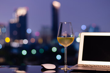 A glass with white wine puts on table with laptop computer and colorful city bokeh lights. Working from home concept.