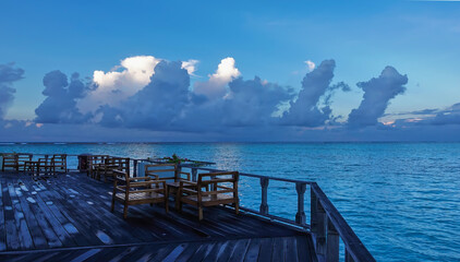 Obraz premium Early morning. On the terrace above the aquamarine ocean, there are tables and chairs damp from the rain. There are picturesque white and blue clouds in the azure sky. Maldivian idyll.
