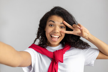 Young beautiful happy African American woman cheerfully smiling and showing V sign while taking selfie on gray background