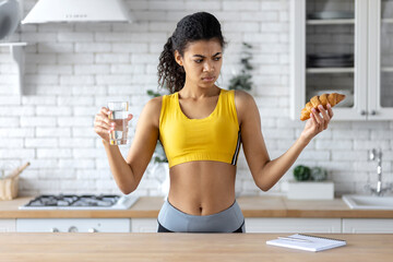 Beautiful young woman fitness trainer holding glass of water and croissant while standing at home kitchen. Start healthy lifestyle or avoiding unhealthy food concept