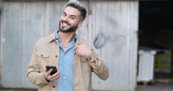 attractive bearded man in denim shirt holding mobile and reading emails, making a call gesture and laughing against grey metal garage outside