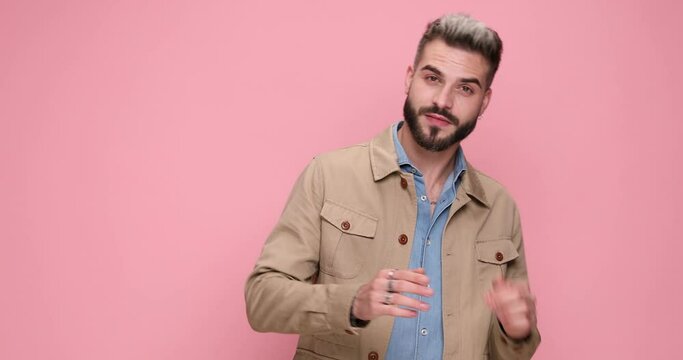 confident cool man in beige jacket rubbing fingers and smiling, making gun and shooting gesture on pink background in studio