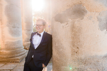 Obraz premium Front view of smiled young male in glasses, wearing in black suit, relying on column of builld, putting hand into pocket of pants and looking away