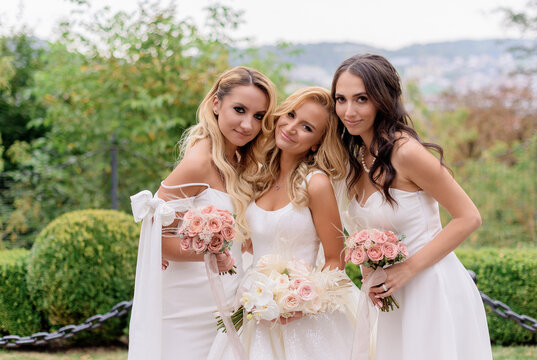 Close of smiled bride, wearing in wedding dress, holding bouquet of roses; standing between bridesmaid and looking at camera
