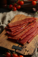 Delicious smoked sausage on a dark wooden board with tomatoes and spices. Delicious handmade snack made from natural ingredients. Pieces of dry pork, beef, chicken snacks. Meat products. Close-up.