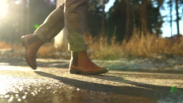 Women legs hiking asphalt . Feet in brown chelsea boots walking on road with sunflares. Girl feet tracking on sunny day outdoors, walking through city park. Middle slow motion tracking shot.