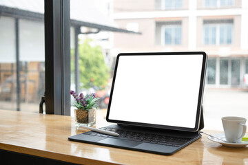 Close up view of computer tablet with white screen, plant and coffee cup on wooden table near window.