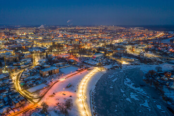 Beautiful evening top view of the city. Evening, night illumination in the city. Winter city in the snow. The river is covered with ice.