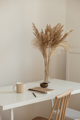 Aesthetic minimal office workspace interior design. Mug, notebook, pampas grass floral bouquet on white table against white wall. Girl, woman boss work at home business concept.
