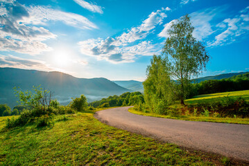 mountain road near meadows and trees on a background of mountains at sunrise. Beautiful summer landscape of mountains.