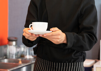 Waiter in black apron holding and serving a cup of hot coffee while standing in coffee bar counter.