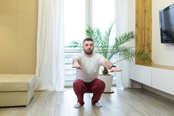 young adult bearded man doing physical exercises stretching at home in the living room or apartment. Male is engaged in fitness and sports workout in the morning sitting on the floor weight loss