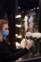 Florist works with colors. Flower seller chooses flowers for future bouquet. Flowers shop worker in a mask standing in flower shop and checking flowers in glass vase.