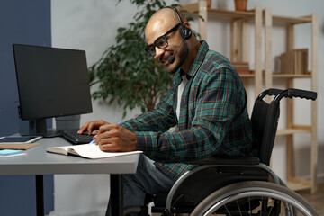 Disabled young man in wheelchair working at his working table with headset on