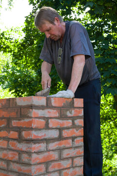 Senior man is making a chimney of red bricks, working with a trowel and making masonry works on a roof of a house, vertical

