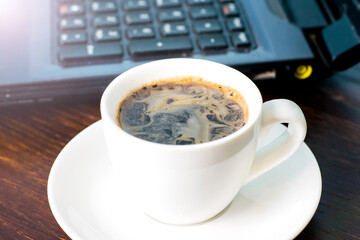 Coffee in a white cup and a laptop on the background on a brown wooden table. Concept of remote work or training, the start of the working day in the office.