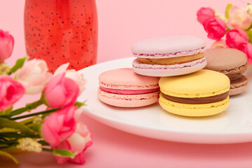 Fototapeta na wymiar Plate of macaroons on pink background with flowers. Drink in bottle. Sweet pastry, baked products, sweets, dessert. Unhealthy diet.