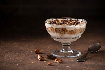 Dessert with yoghurt cream, chocolate and nuts in a glass bowl on a dark brown stone background with blank space on the left