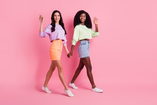 Full length body size view of attractive cheerful girls going holding hands waving hi isolated over pink pastel color background
