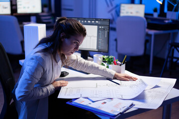 Overworked woman architect checking and matching blueprints sitting at office desk in front of...