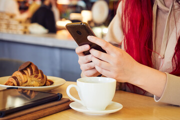 Hands of a woman with red hair is typing a message in the phone while sitting in a restaurant over a cup of coffee with a croissant.