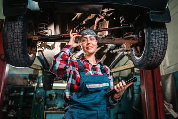 Fototapeta na wymiar A young pretty smiling female mechanic, in a uniform and glasses, with a tablet in her hands, poses standing under a car on a lift. Indoors garage