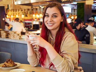 Pretty young woman with red hair enjoying a cup of coffee with a croissant in a restaurant.
