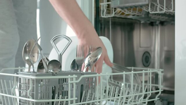 A dishwasher is built into the white kitchen unit. Open the dishwasher door, slide out the bottom drawer with kitchen utensils. The dishes are taken out one by one. Close-up