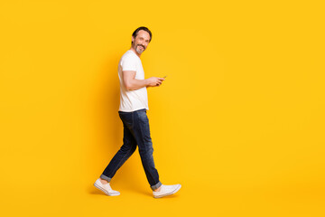 Full body profile side photo of charming happy man walk smile good mood hold smartphone isolated on yellow color background