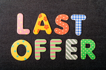 Card with Last Offer words made from mixed vivid colored wooden letters on a textured dark black textile material that can be used as a message.