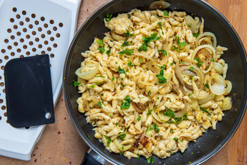 Spaetzle or Spätzle egg noodle pasta with onions, parsley and seasoning in a pan. Traditional European cuisine dish (Swabian, German, Austrian). Handmade gluten free Knöpfle. Grater in background