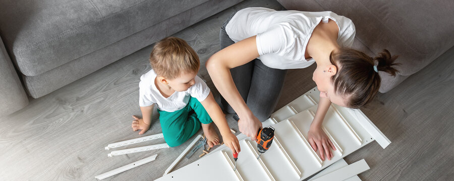 Cute Adorable Caucasian Toddler Boy Kid Sit On Floor And Help Mom Assembling Furniture Shelf With Power Screwdriver Tool. Young Adult Mum With Funny Little Child Enjoy Playing At Home Indoors