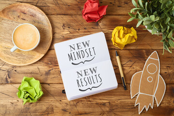 New mindset and new results text on notebook on wooden table with coffee cup and paper rocket. Business concept of positive thinking and motivation