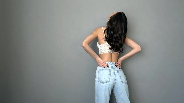 pretty young brunette girl in blue jeans and white bra standing back to the camera posing on the grey background copy-space 