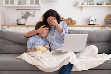 Frightened mom and little kid son with bowl of popcorn watching scary scene in movie closing their eyes, sitting on sofa at home. Mother hugs the child, watches a horror movie, feels fear.