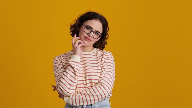 A displeased young teenager girl in eyeglasses is looking to the camera standing isolated over yellow background in studio