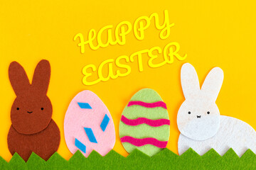 Cut out of felt applications of two eggs and white and brown rabbits on the grass. Yellow background. Flat lay. Easter holiday concept